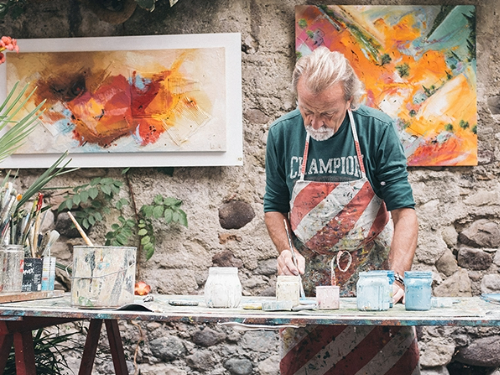 An artist wearing a paint covered apron painting whilst stood over a table full of art supplies.