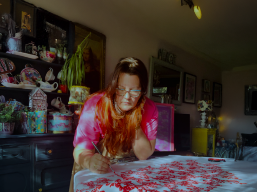 A lady sat at a table painting red flowers over a white tablecloth.