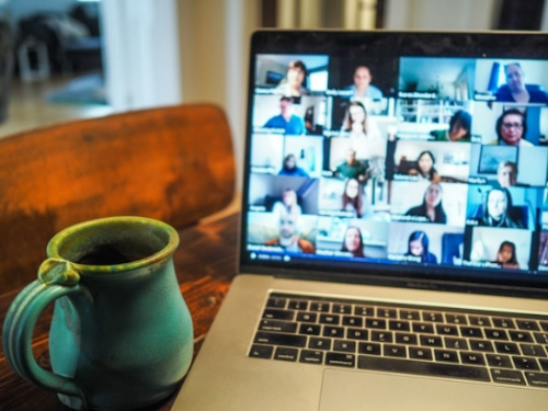 An image of a mug next to a laptop showing a Zoom conference call with many peoples webcams open showcasing their faces.