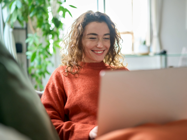 Smiling woman in orange jump sat on sofa with laptop on knees.